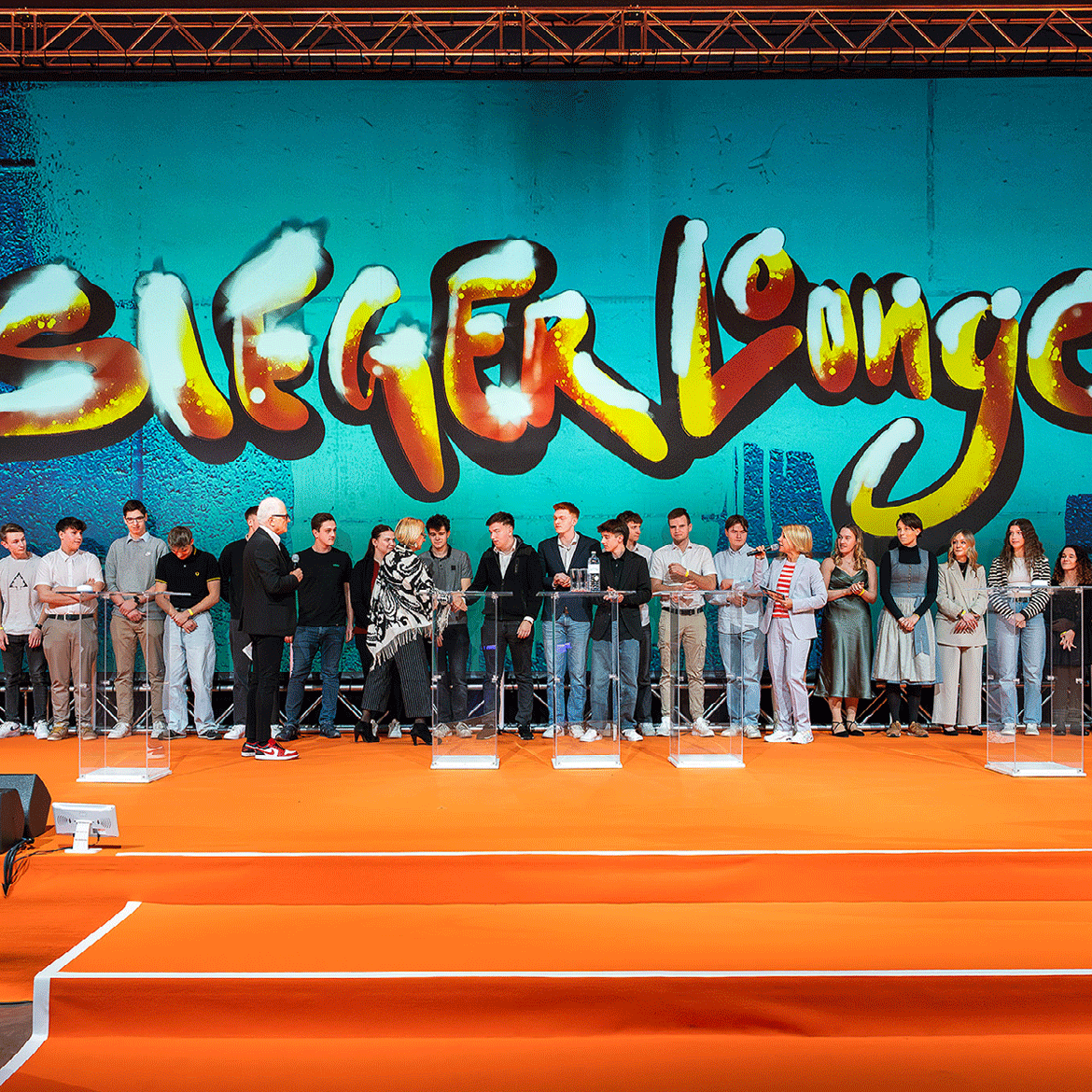 Eine Linie von Menschen auf einer Bühne, im Hintergrund steht Siegerlounge an der Wand.