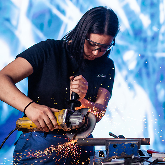 Mädchen in Schutzkleidung schneidet ein Metallstück mit einer Kreissäge zu. Die Funken sprühen.