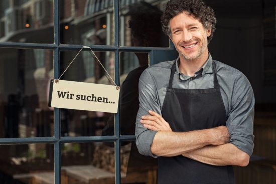 Ein Mann vor einem Schaufenster. Daneben ein Schild mit dem Text: Wir suchen