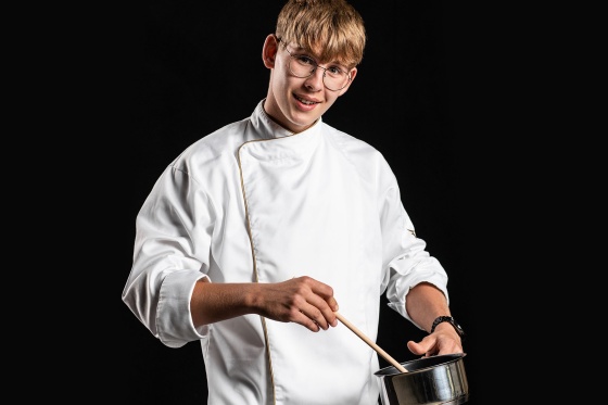 Ein Junge in Kochkleidung hält Topf, Kochlöffel und lächelt in die Kamera.