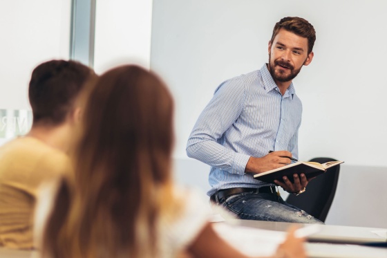 Ein junger Lehrer lehnt auf einem Pult und blickt auf verschwommen sichtbare Schüler.