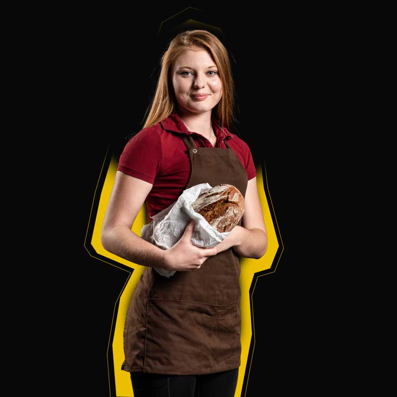 Eine junge Bäckerin mit Brot in der Hand steht vor einem schwarzen Hintergrund.